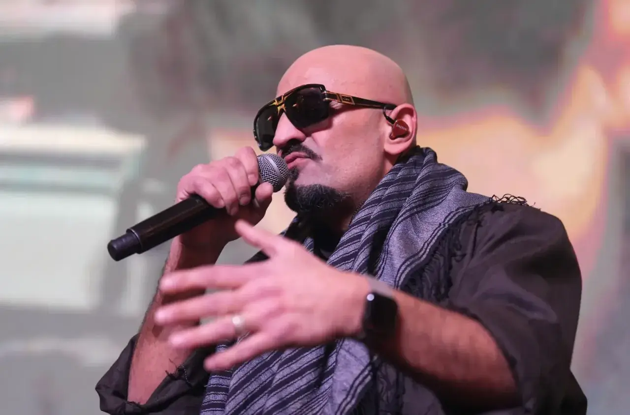 Ein bekannter Rapper mit Sonnenbrille und Schal spricht ins Mikrofon, während er gestikuliert.