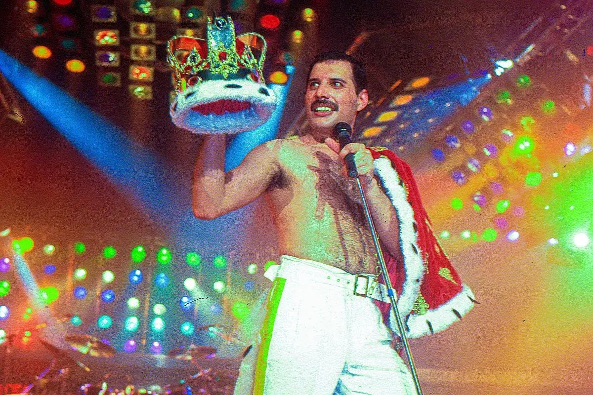 Freddie Mercury, w koronie i z mikrofonem, na scenie pełnej świateł. Pamięć o nim żyje, choć zmarł Freddie Mercury.