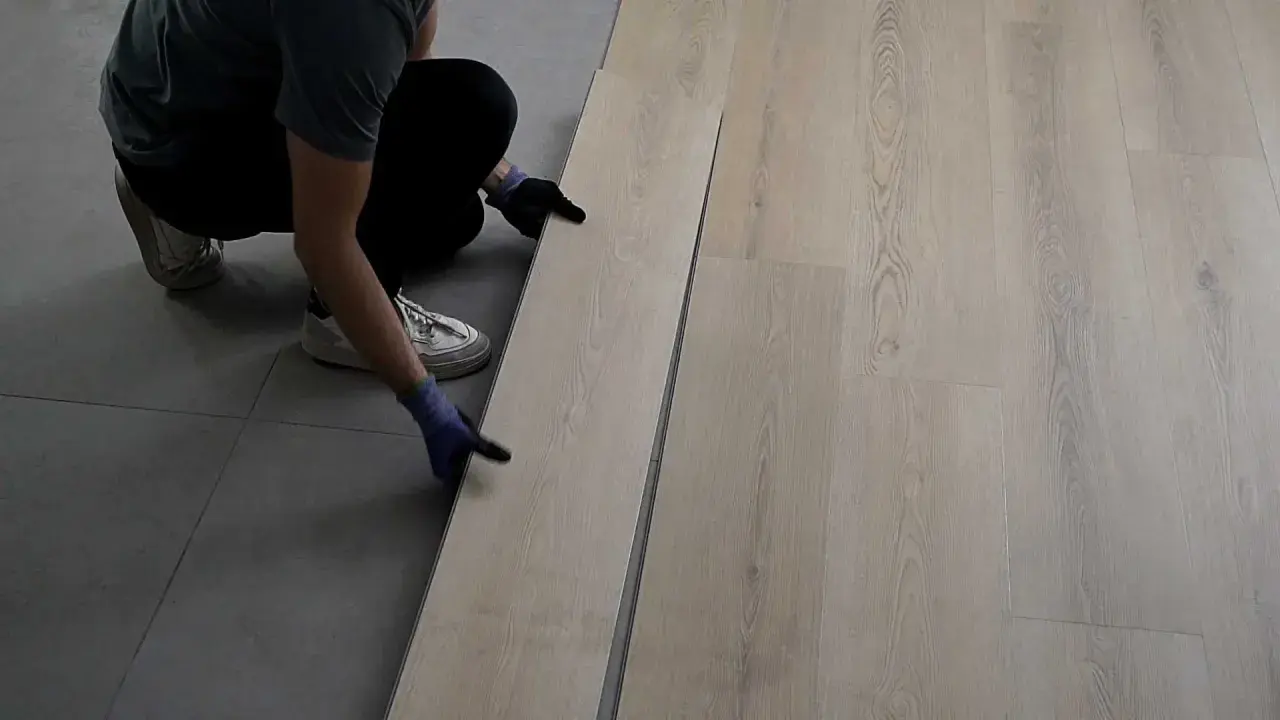 Persona con guantes instala piso SPC de madera clara sobre suelo gris.