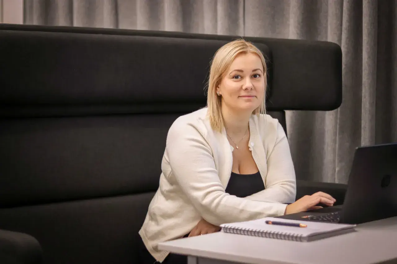 Kvinna med blont hår sitter vid ett bord med laptop och anteckningsblock. Hon har en vit kofta över en svart topp. Möjlighet till gratis webbutbildning med diplom.
