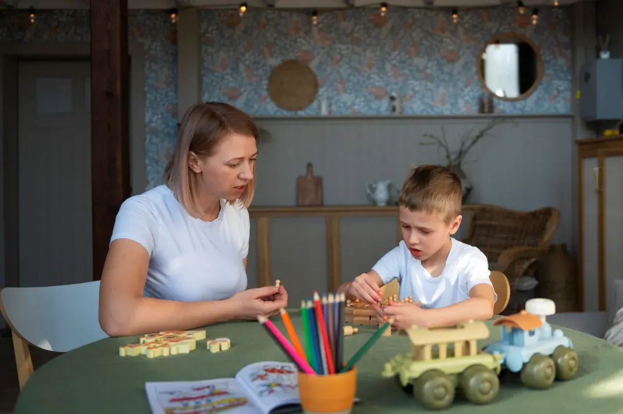Kobieta i chłopiec grają w drewniane gry na zielonym stole. W tle widać wystrój pokoju, przypominający salę w przedszkolu niepublicznym.