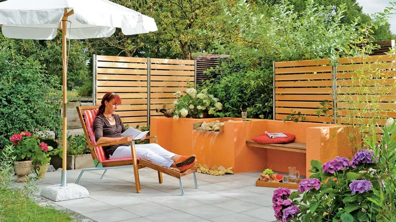 Entspannter Garten mit selbst gebautem Wasserfall. Eine Frau liest unter einem Sonnenschirm.