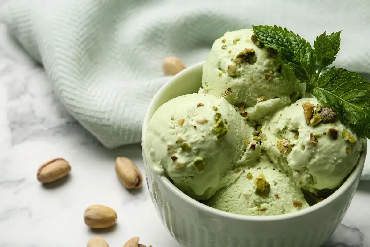 Coppa di cremoso gelato al pistacchio, guarnita con foglioline di menta e granella di pistacchi. Alcuni pistacchi interi sparsi sul marmo.