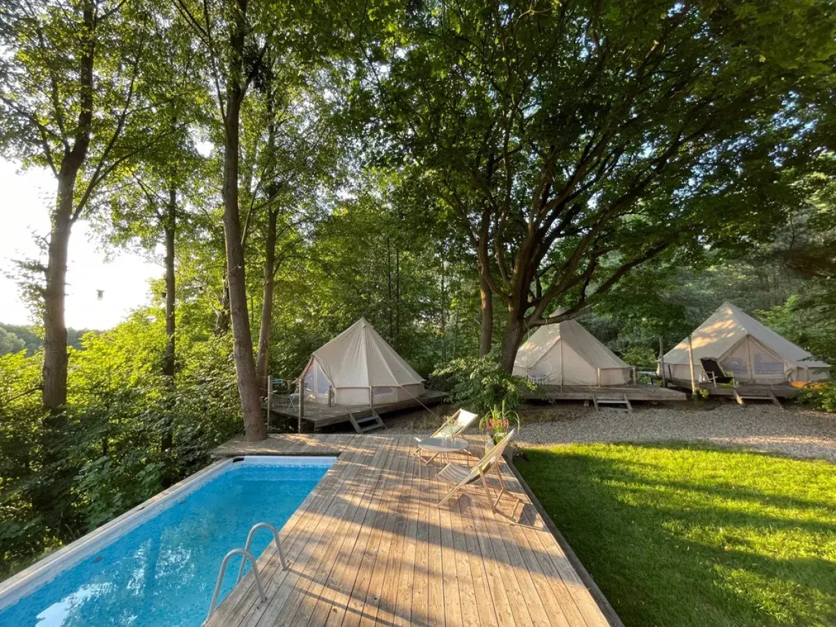 Luksusowy glamping nad jeziorem. Białe namioty na drewnianych tarasach, basen i leżaki wśród zieleni.