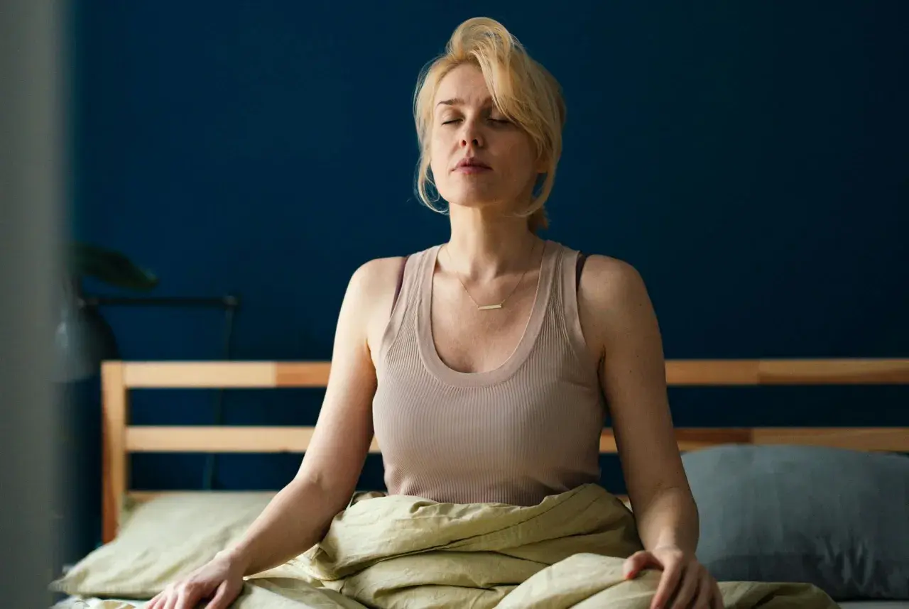 Kvinna med slutna ögon sitter i sängen, insvept i täcke, i en avslappnad somna meditation.