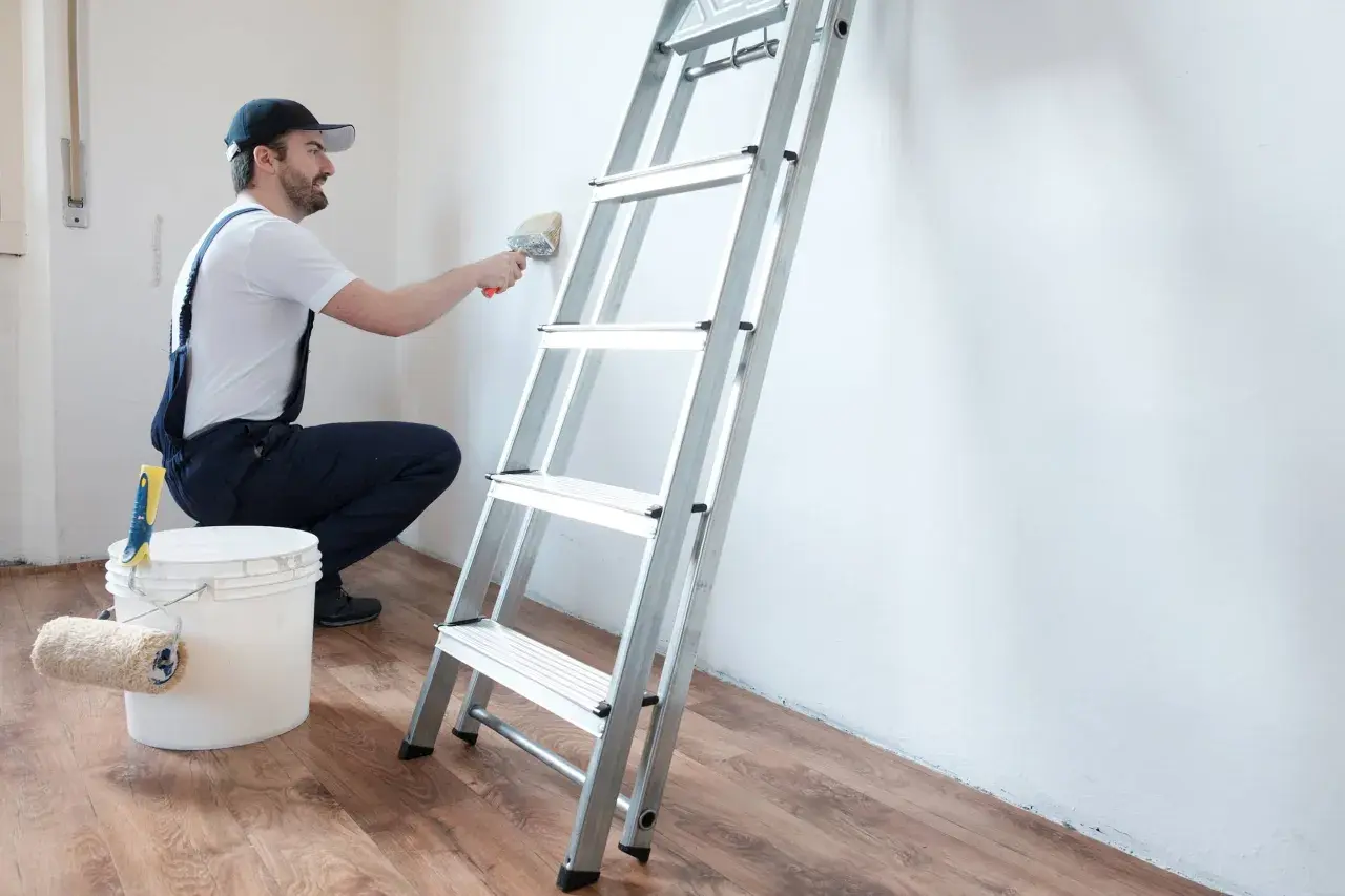 Hombre con brocha y cubo de pintura, listo para limpiar pared antes de pintar. Escalera metálica al lado.