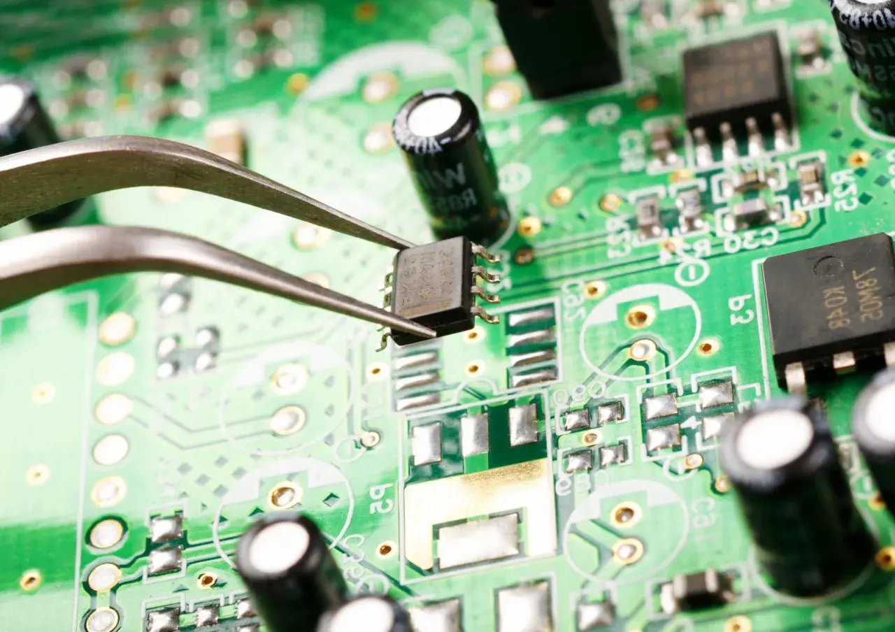 Pęseta trzyma układ scalony na zielonej płytce drukowanej. Widać elementy elektroniczne, kondensatory i ścieżki. Zbliżenie ujawnia potencjalne oznaki korozji elektrochemicznej.