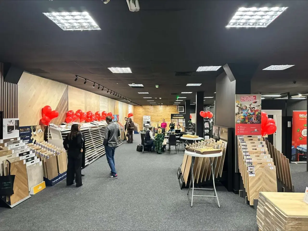 Wnętrze salonu z wystawionymi panelami podłogowymi, dekoracjami z balonów i ludźmi.