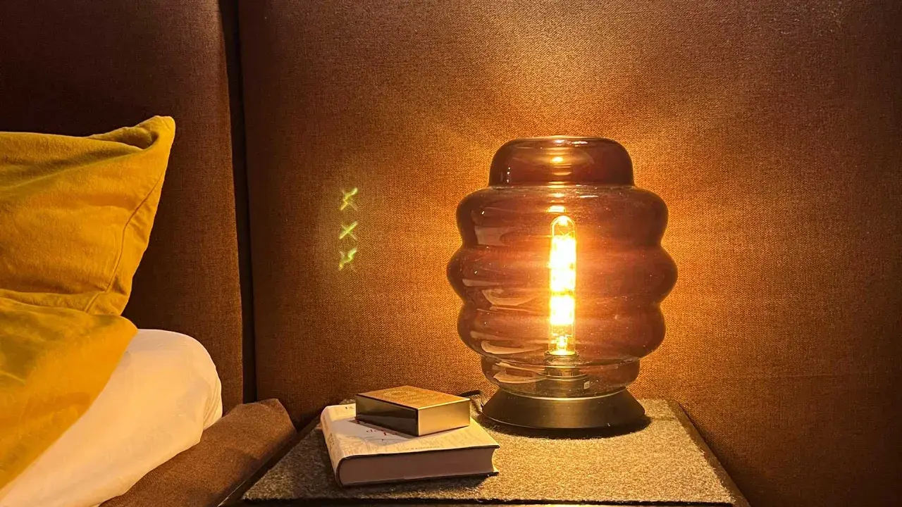 Una lámpara de mesa de cristal ahumado ilumina un rincón acogedor junto a una cama con almohada amarilla.