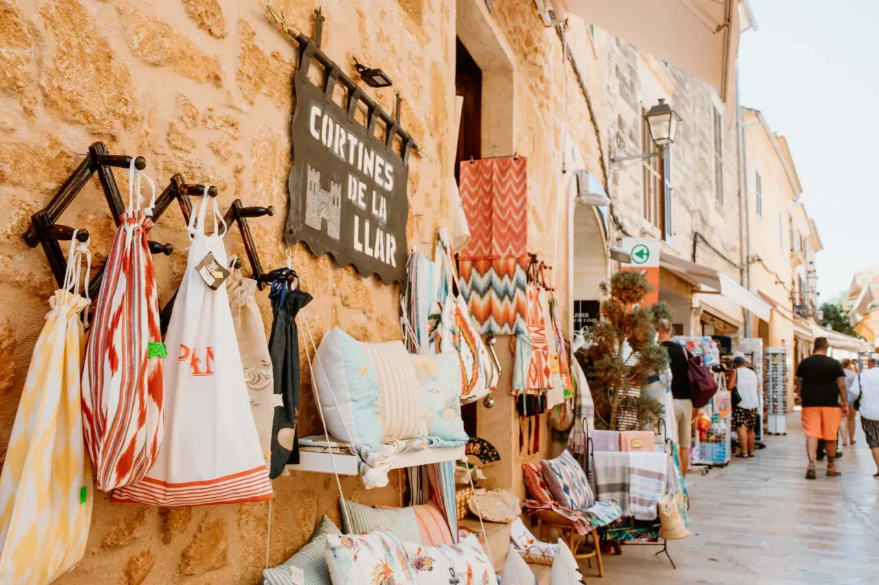 Bunte Stoffe und Kissen hängen vor einem Geschäft am alcudia markt. Menschen schlendern die Gasse entlang.
