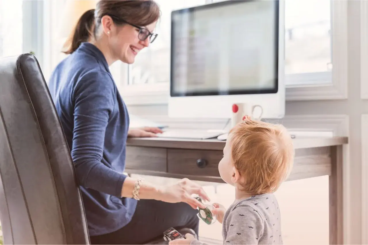 En kvinna med glasögon ler mot sitt barn vid ett skrivbord. Hon visar vad betyder flexibel genom att balansera arbete och familjeliv.