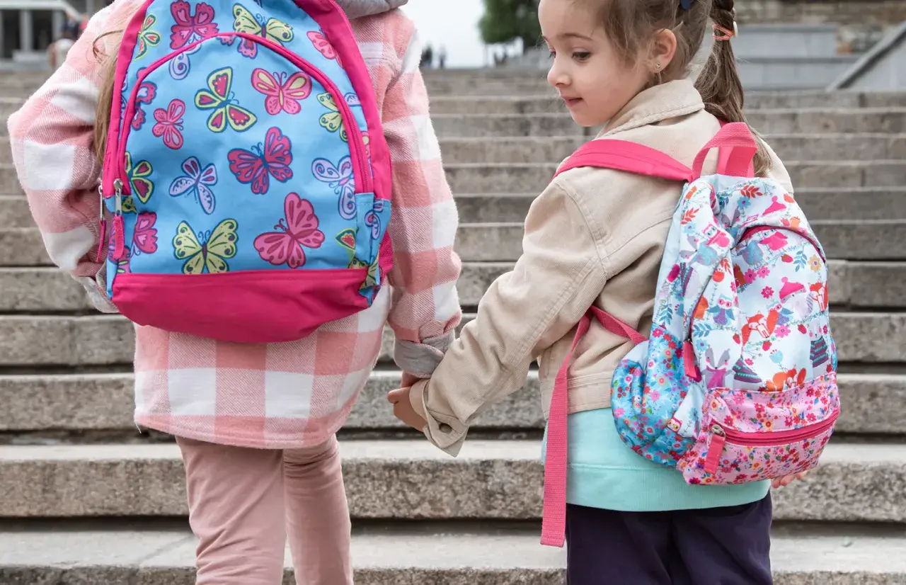 Dwie dziewczynki w kolorowych plecakach szkolnych trzymają się za ręce na schodach.