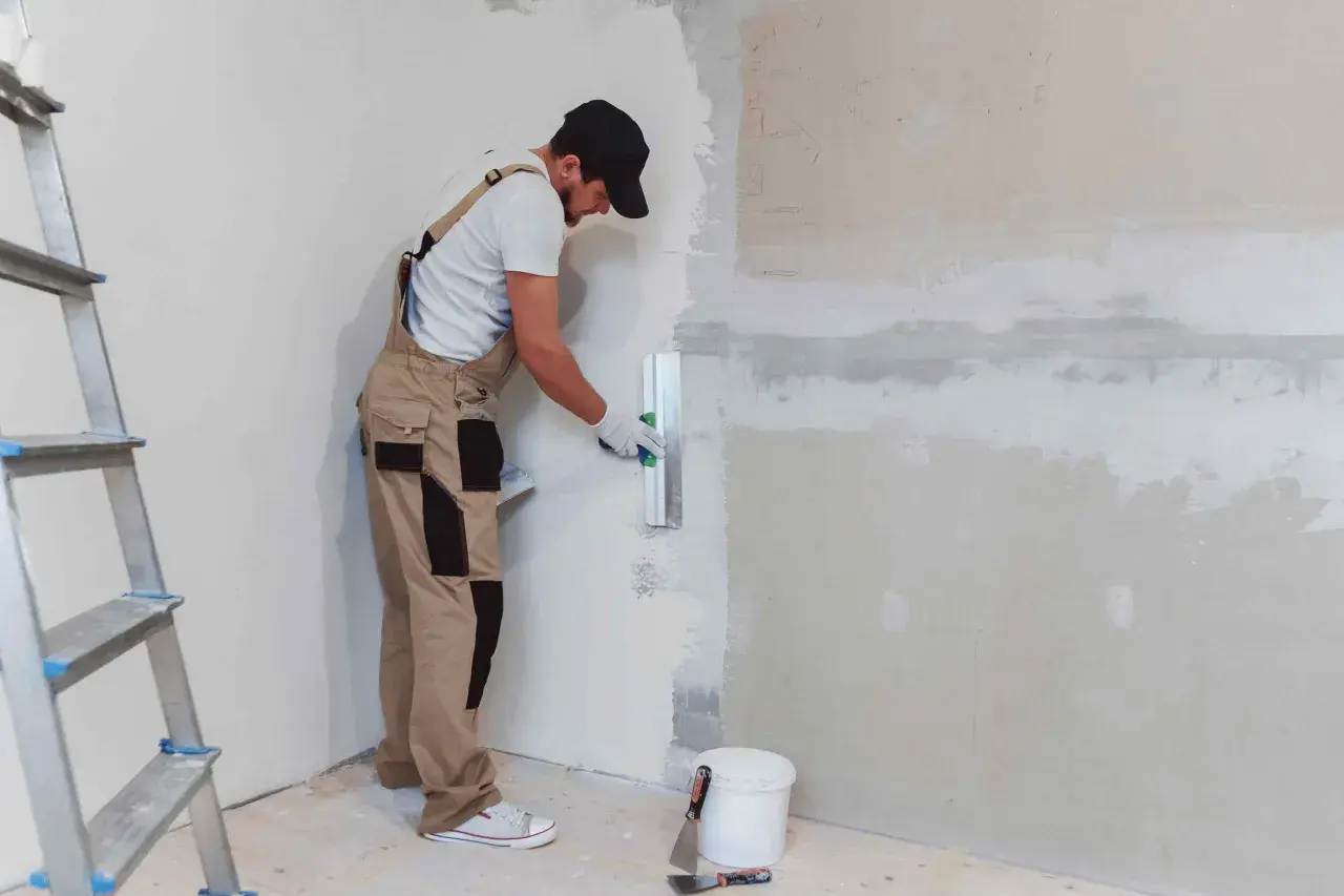 Operaio con tuta beige e cappellino nero mentre impara come intonacare un muro, usando una spatola. Scala e secchio a terra.