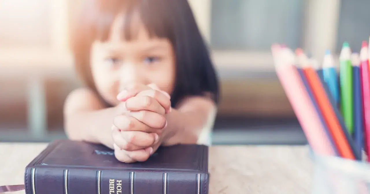 Dziecko z modlącymi się dłońmi na Biblii, obok kolorowe kredki. Nauka 10 przykazań bożych dla dzieci.