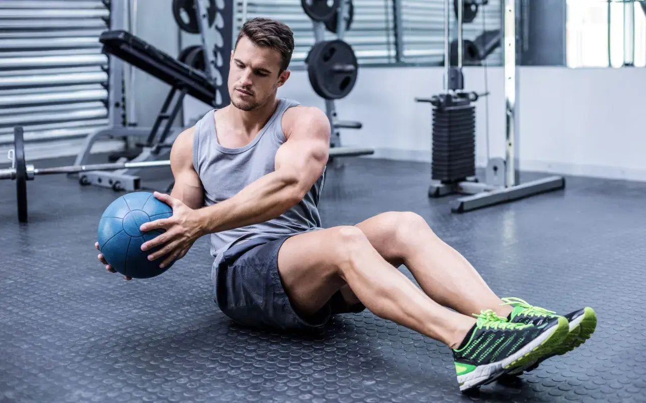 Hombre sentado en el suelo del gimnasio, girando el torso con una pelota medicinal azul, trabajando la elasticidad muscular.