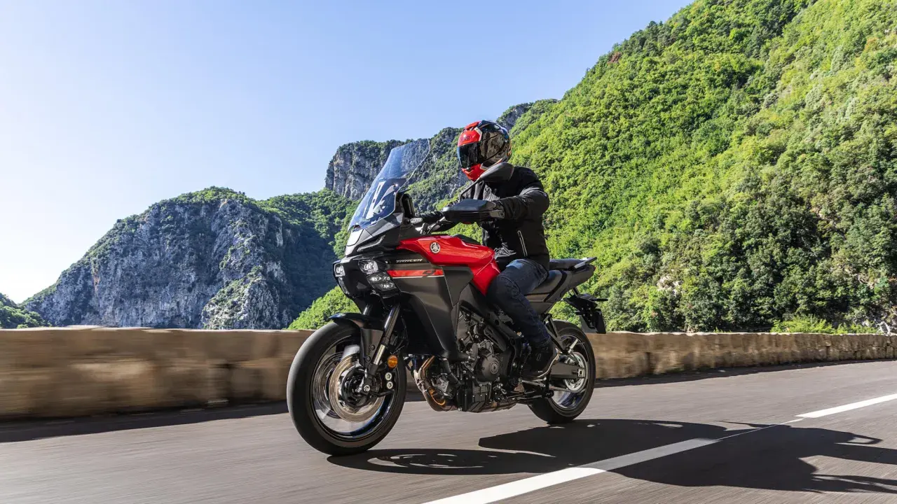 Un motociclista su una Yamaha Tracer rossa affronta una curva su una strada panoramica, godendosi una gita in moto tra montagne verdi e cielo azzurro.