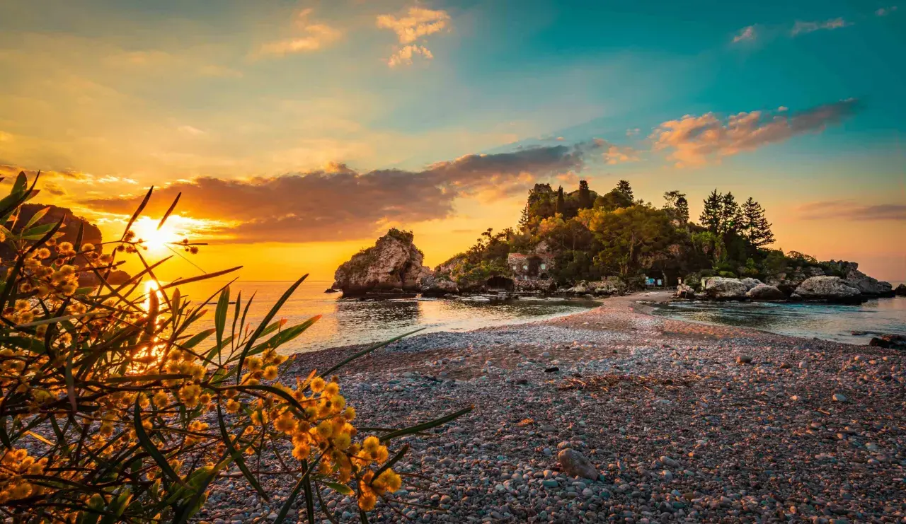 Tramonto dorato sulle spiagge di Giardini Naxos Taormina, con rocce e vegetazione lussureggiante.