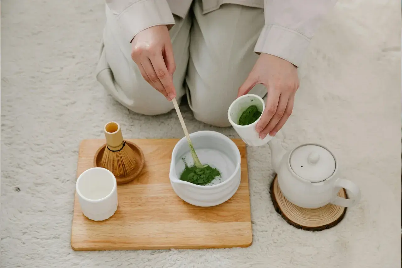 Hände bereiten Matcha zu. Ein Bambusbesen und eine Tasse stehen bereit. Dieser Matcha macht wach!