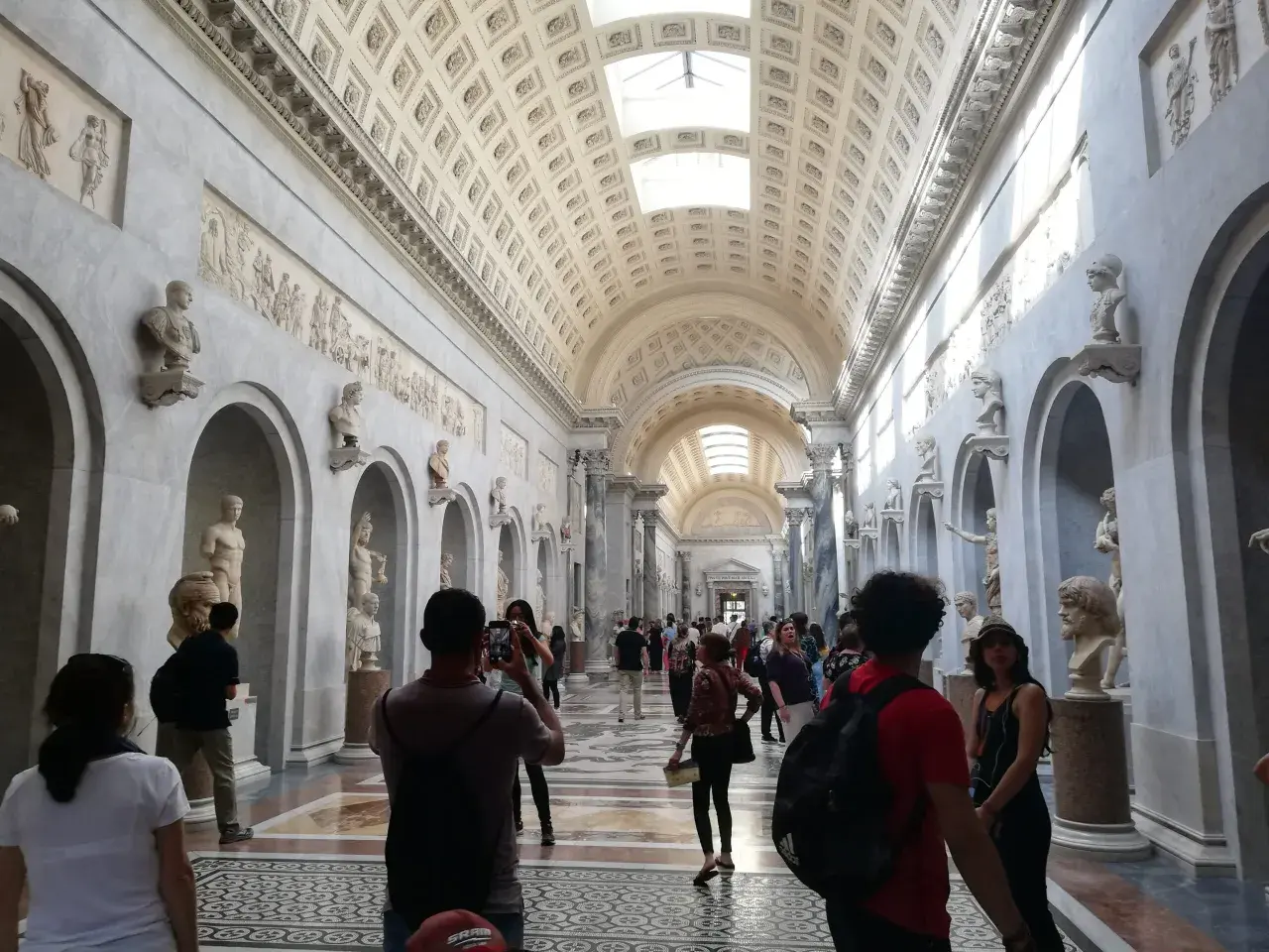 Visiteurs admirant des sculptures dans un long couloir orné de bustes et de bas-reliefs. Une excellente façon de savoir comment aller au Vatican.