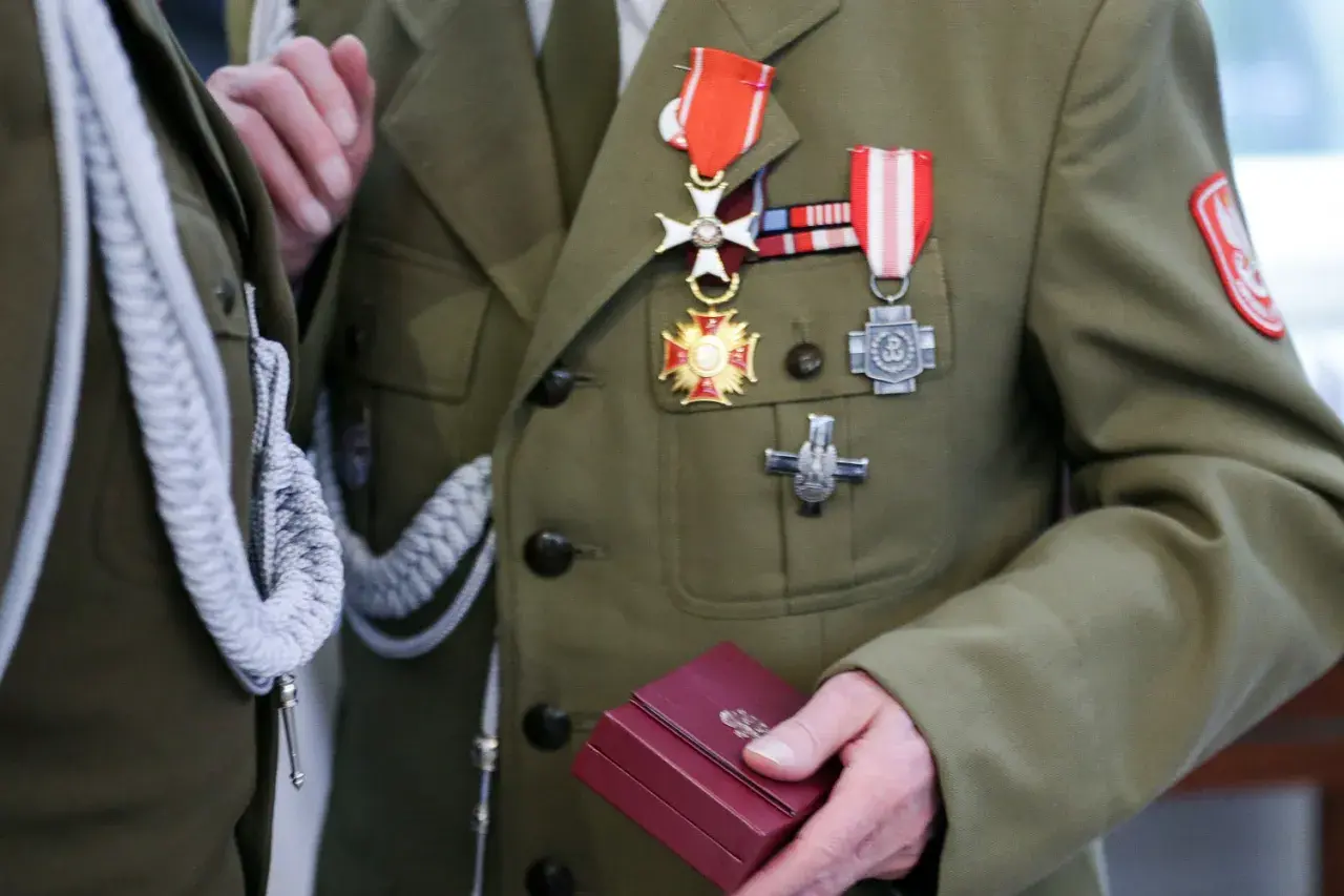 Żołnierz w mundurze z licznymi odznaczeniami wojskowymi na piersi. W dłoni trzyma małe, czerwone pudełko.
