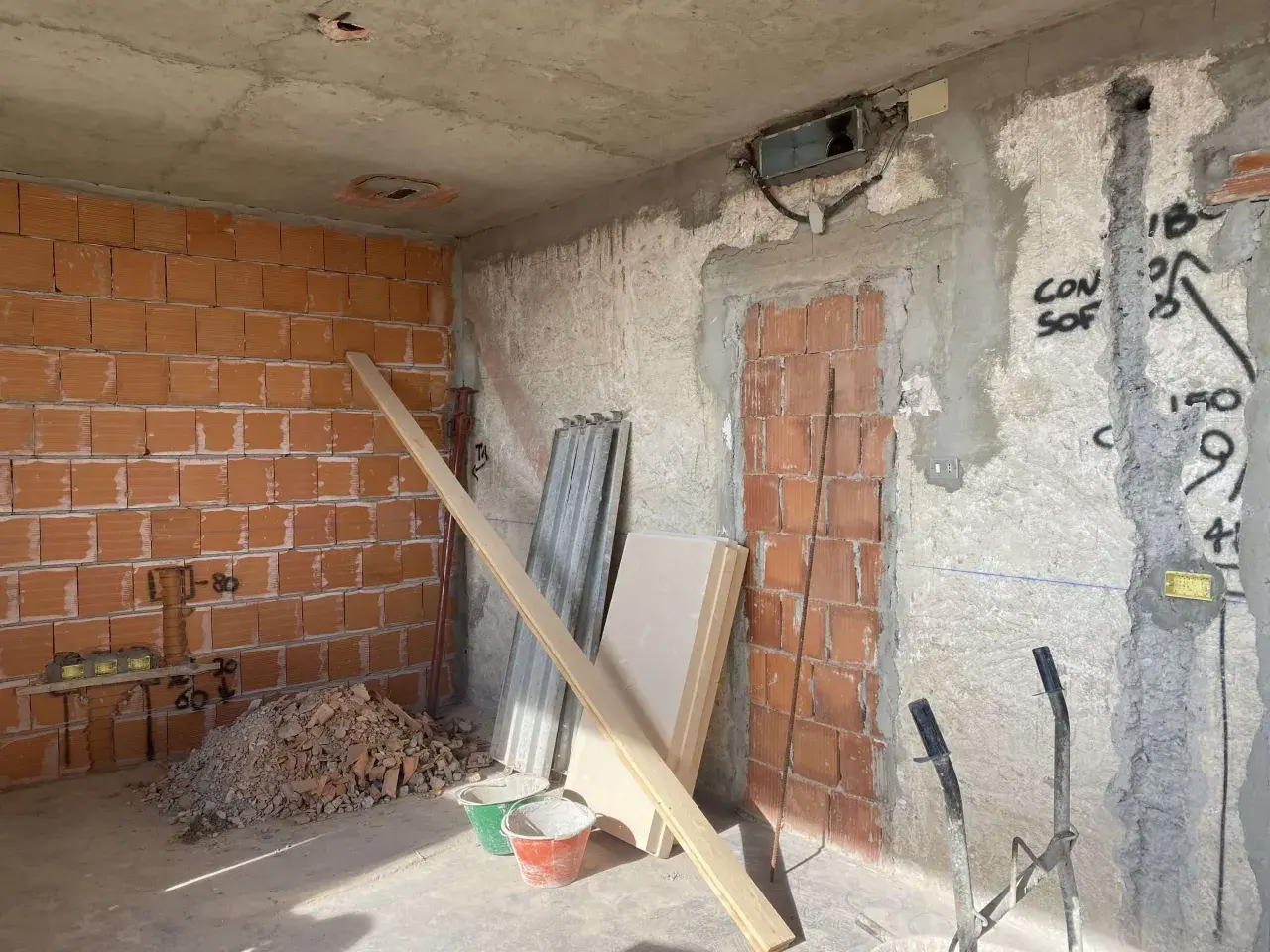 Interno di una casa in fase di ristrutturazione: pareti in mattoni a vista, cumulo di macerie, secchi, assi di legno e materiale edile.