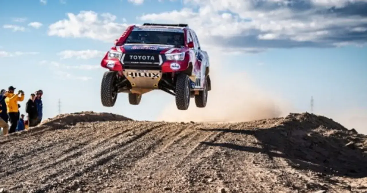 Toyota Hilux w locie podczas rajdu Dakar. Widzowie obserwują zmagania na pustynnym terenie.