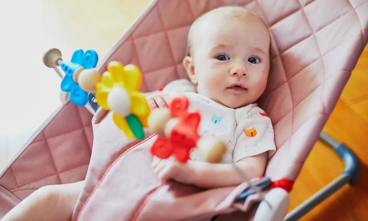 Söt bebis i en rosa babysitter vagga med färgglada leksaker.