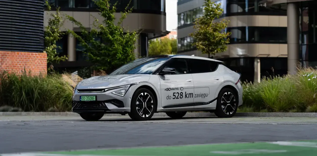 Biały Kia EV6 zaparkowany przed nowoczesnym budynkiem, z napisem "#GOelectric do 528 km zasięgu".