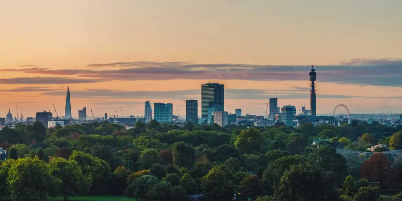 London im Sommer: Die Skyline mit dem Shard und dem BT Tower ragt über einem grünen Park auf, während die Sonne den Himmel in warme Farben taucht.