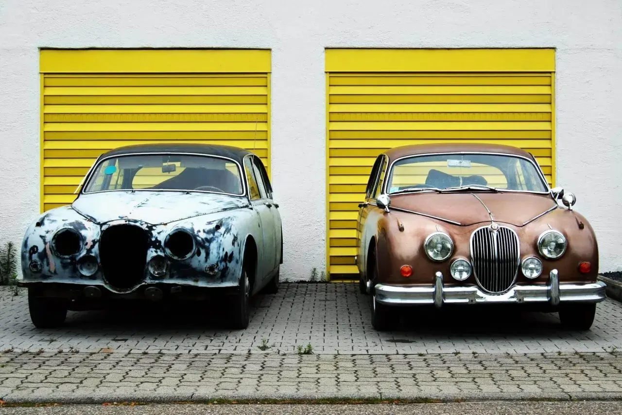 Zwei Oldtimer vor gelben Garagentoren. Einer ist stark verrostet, der andere glänzt. Überlegen Sie, ob auto selber lackieren kosten günstiger sind.