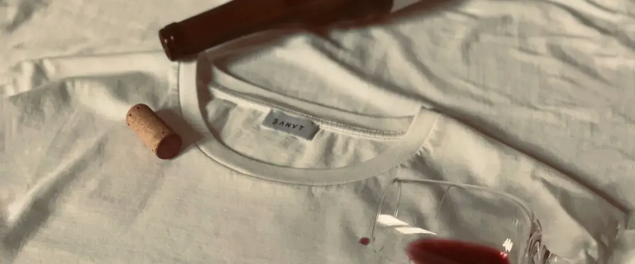 Un bouchon de liège et une bouteille de vin à côté d'un tee shirt blanc, avec une tache de vin rouge.