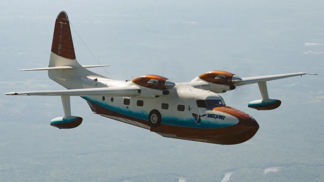 Grumman Mallard Wasserflugzeug fliegt über bewaldetem Gebiet. Das Flugzeug hat eine weiße und braune Lackierung mit blauen Akzenten.