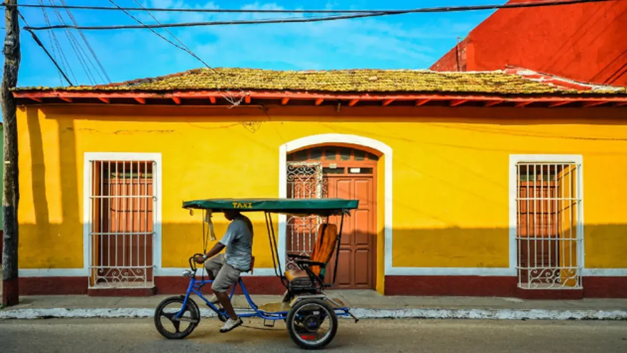 Un hombre pedalea un taxi bicitaxi frente a una casa amarilla vibrante, un recuerdo de mi viaje a Cuba.