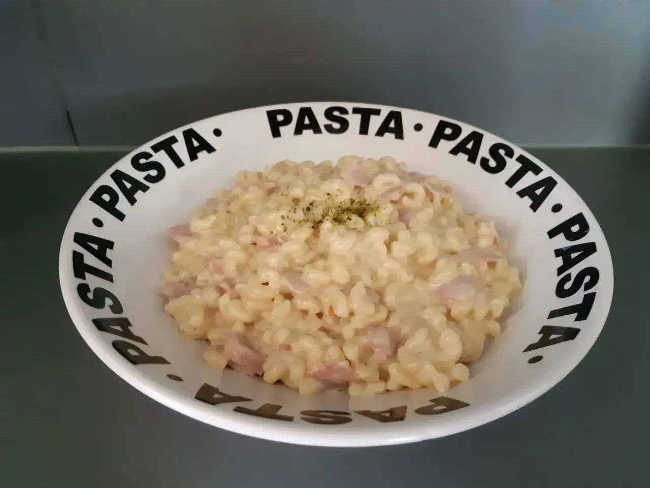 Un crémeux risotto coquillette jambon, garni d'herbes, servi dans une assiette au message "PASTA" répété.