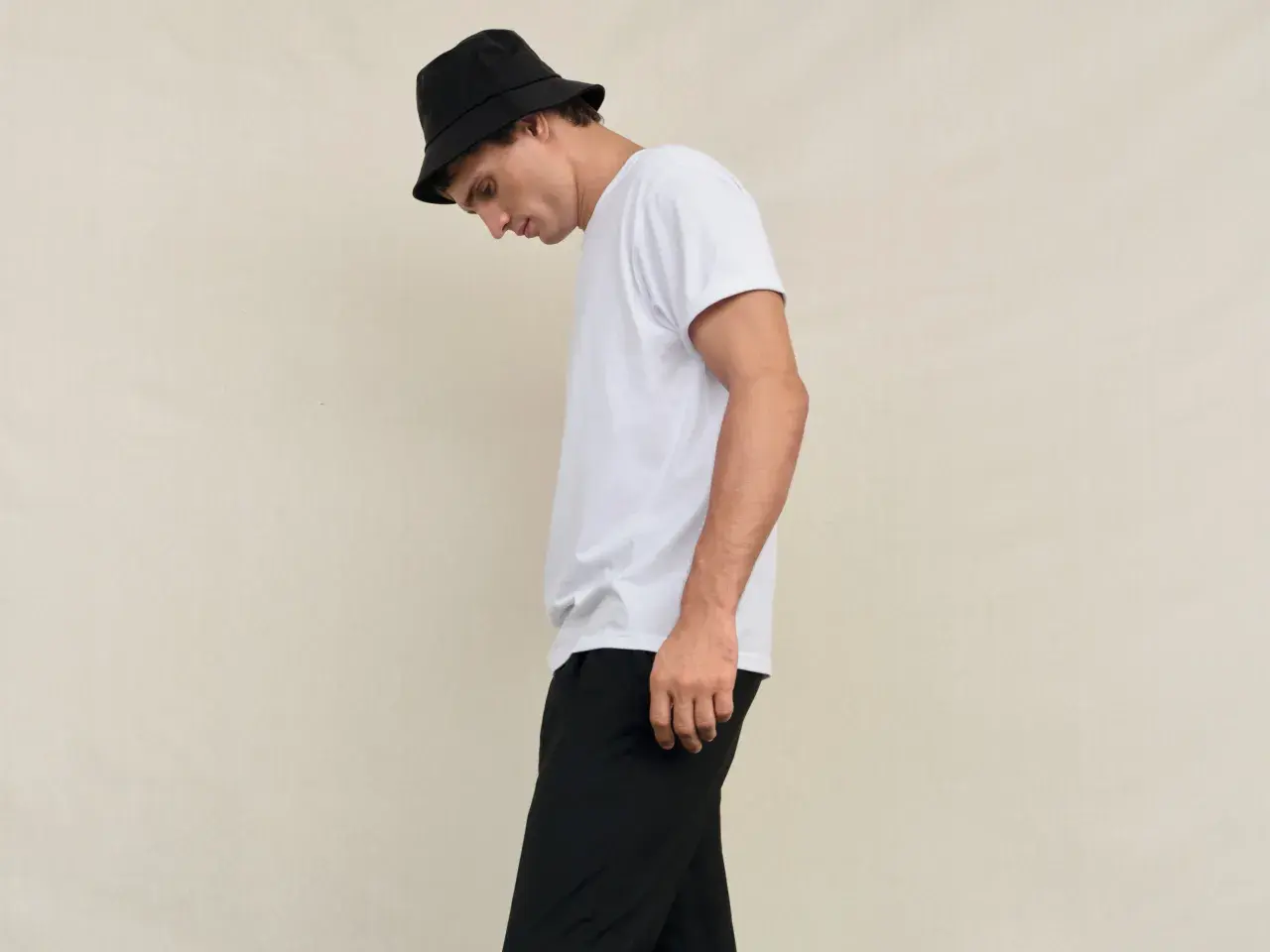 Młody mężczyzna w czarnym kapeluszu typu bucket hat i białej koszulce, spodnie w stylu skate.