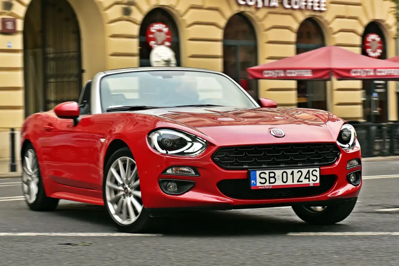 Czerwony Fiat 124 Spider z otwartym dachem sunie ulicą przed kawiarnią Costa Coffee.