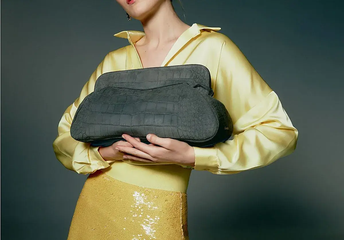 Elegante bolso gris de piel de cocodrilo, un accesorio de lujo de marcas españolas, sostenido por una mujer con blusa amarilla satinada y falda de lentejuelas.