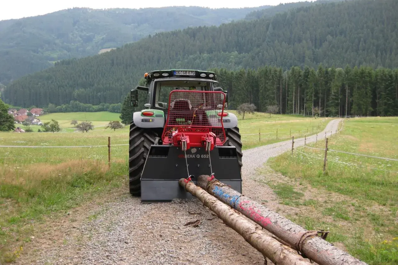 Traktor mit angehängter seilwinde forst zieht zwei Baumstämme über einen Schotterweg, umgeben von grünen Wiesen und einem dichten Wald.