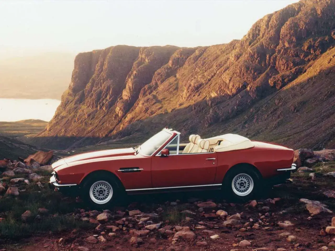 Röd Aston Martin V8 Vantage cabriolet parkerad i en stenig, bergig terräng med en sjö i bakgrunden.