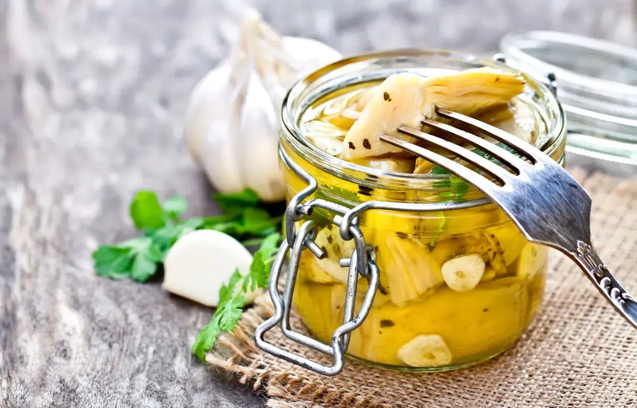 Un barattolo di carciofini sott'olio alla contadina, con aglio e prezzemolo. Una forchetta solleva un carciofino, pronto per essere gustato.