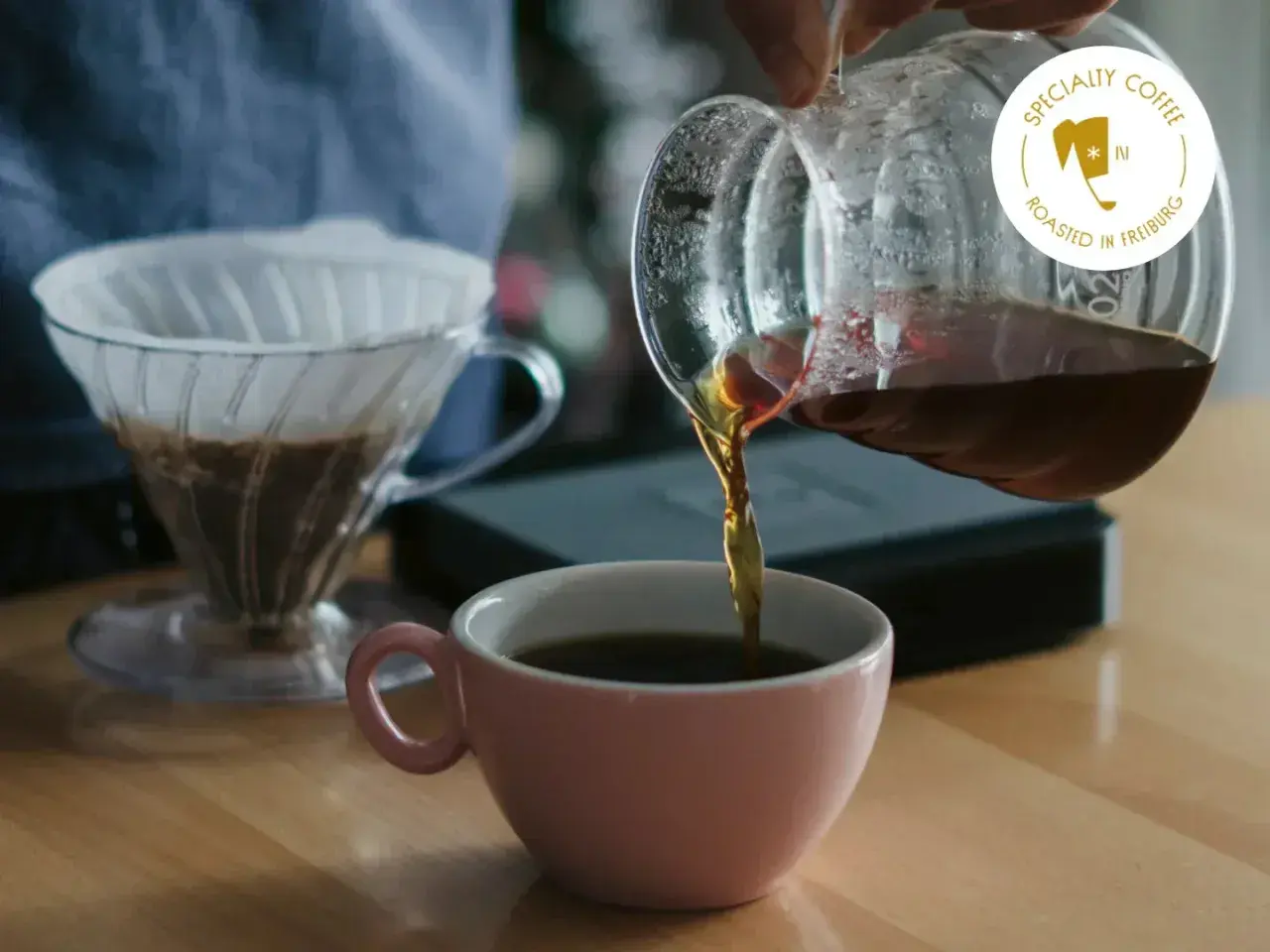 Frisch gebrühter Kaffee fließt aus einer Glaskaraffe in eine rosa Tasse. Ein V60-Filter mit Kaffee steht bereit. Das perfekte Kaffee Rezept für Genießer.