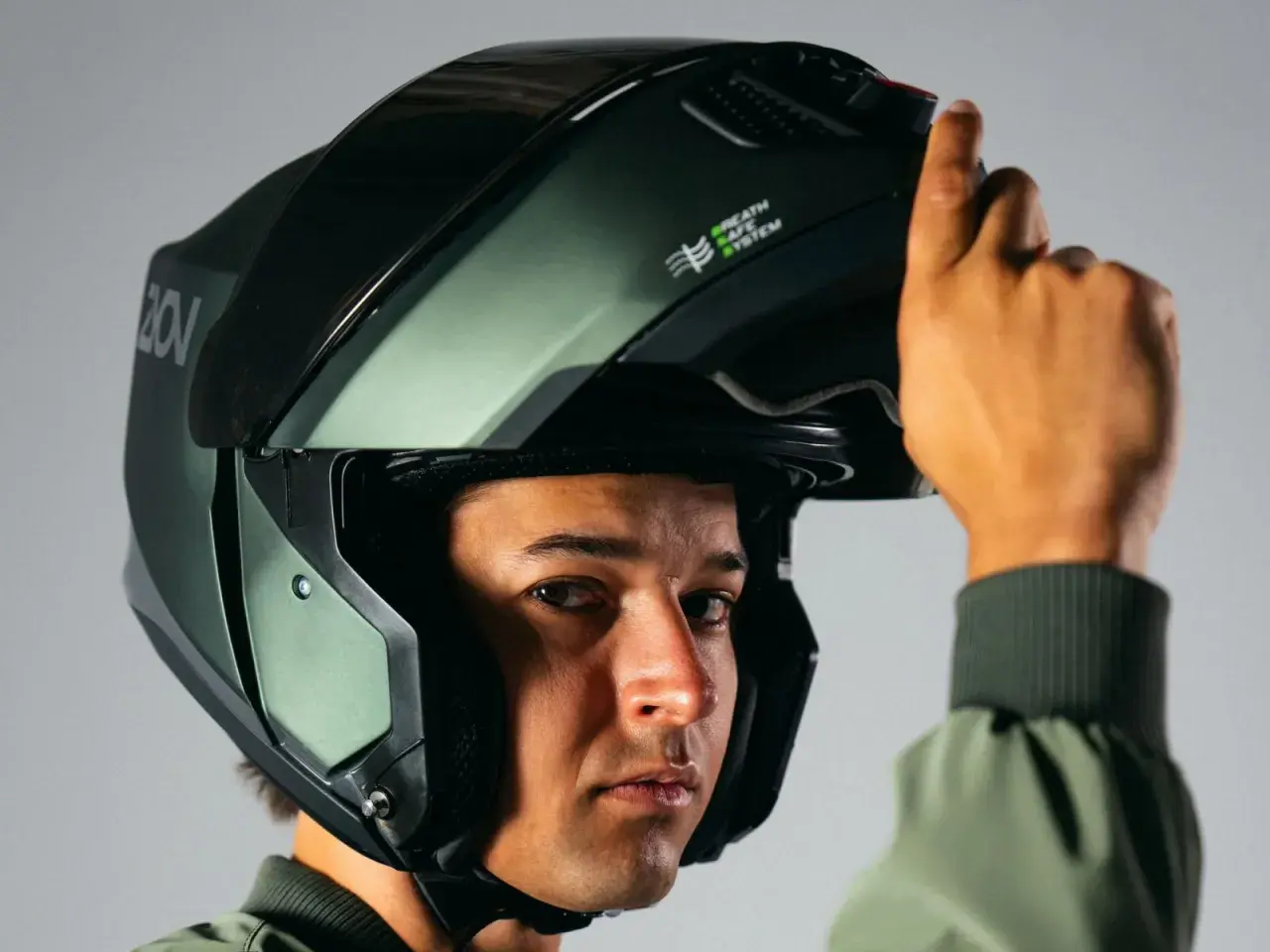 Un hombre se ajusta un casco de moto verde oscuro, similar a un casco guardia civil, con el sistema "Breath Safe" visible.