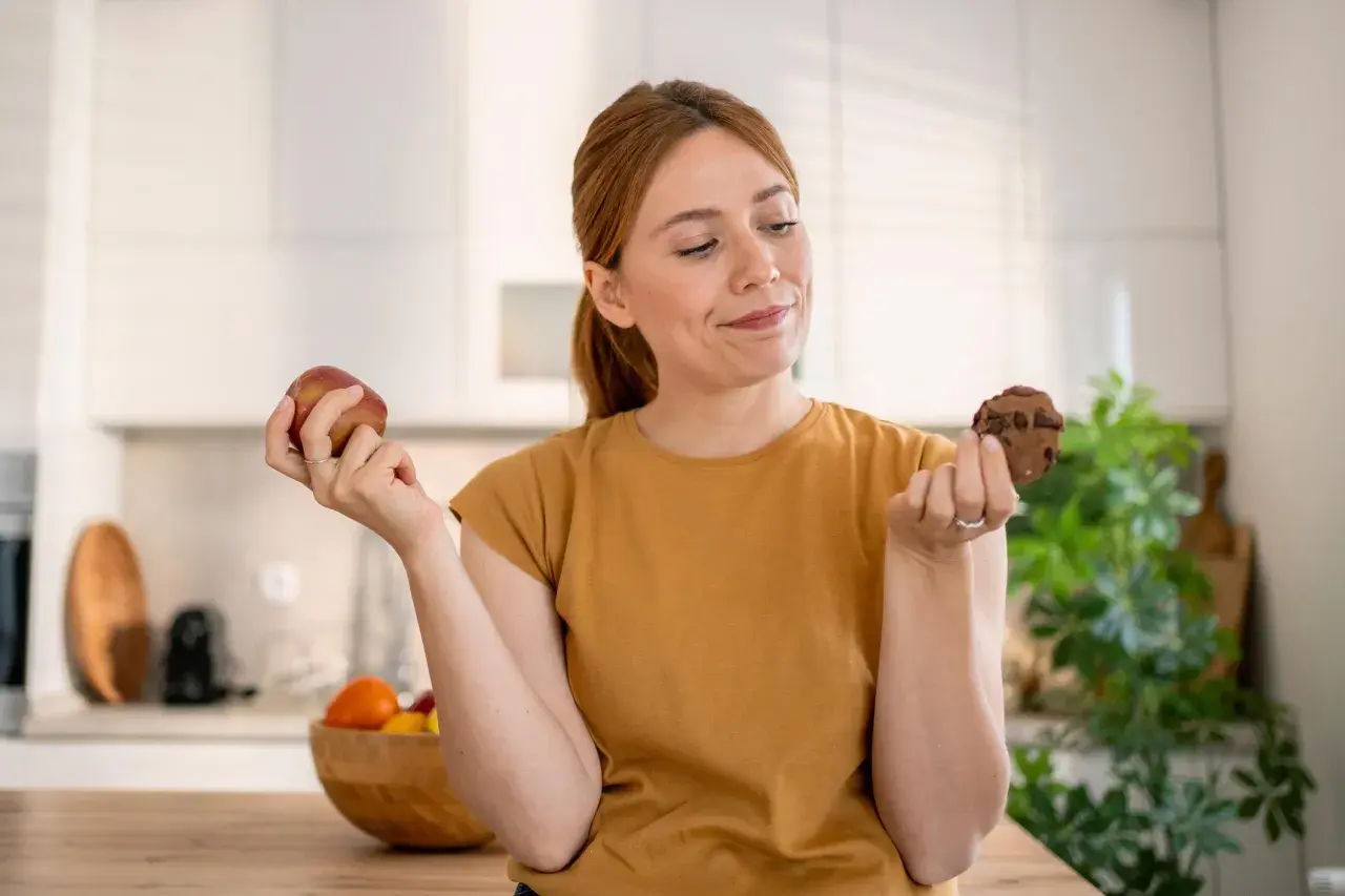 Vrouw twijfelt tussen een appel en een chocoladekoekje. De strijd tegen het verslaafd zijn aan eten is zichtbaar in haar blik.