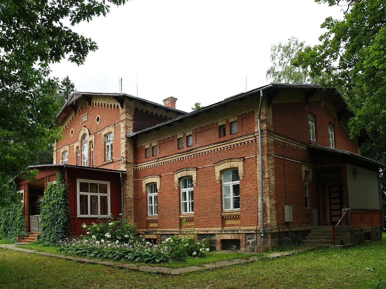 Stary, ceglany maison otoczony zielenią, z werandą i kwitnącymi krzewami.