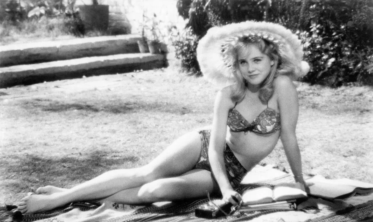 Młoda kobieta w bikini i kapeluszu leży na trawie, czytając książkę. Scena jak z letnich filmów erotycznych.
