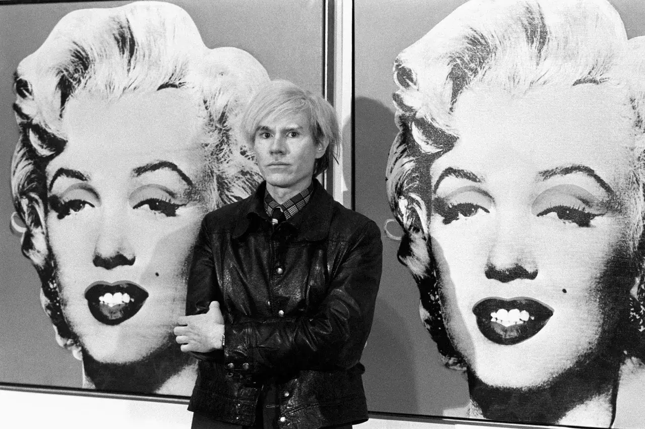Andy Warhol posa tra i suoi iconici ritratti di Marilyn Monroe, un'immagine che definisce la Pop Art.