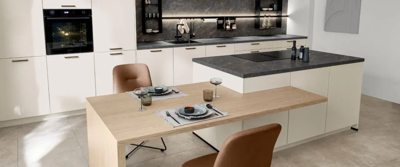 Cocina moderna con encimeras de piedra oscura y gabinetes blancos. La mesa de madera clara y las sillas de cuero marrón complementan los materiales para cocinas modernas.