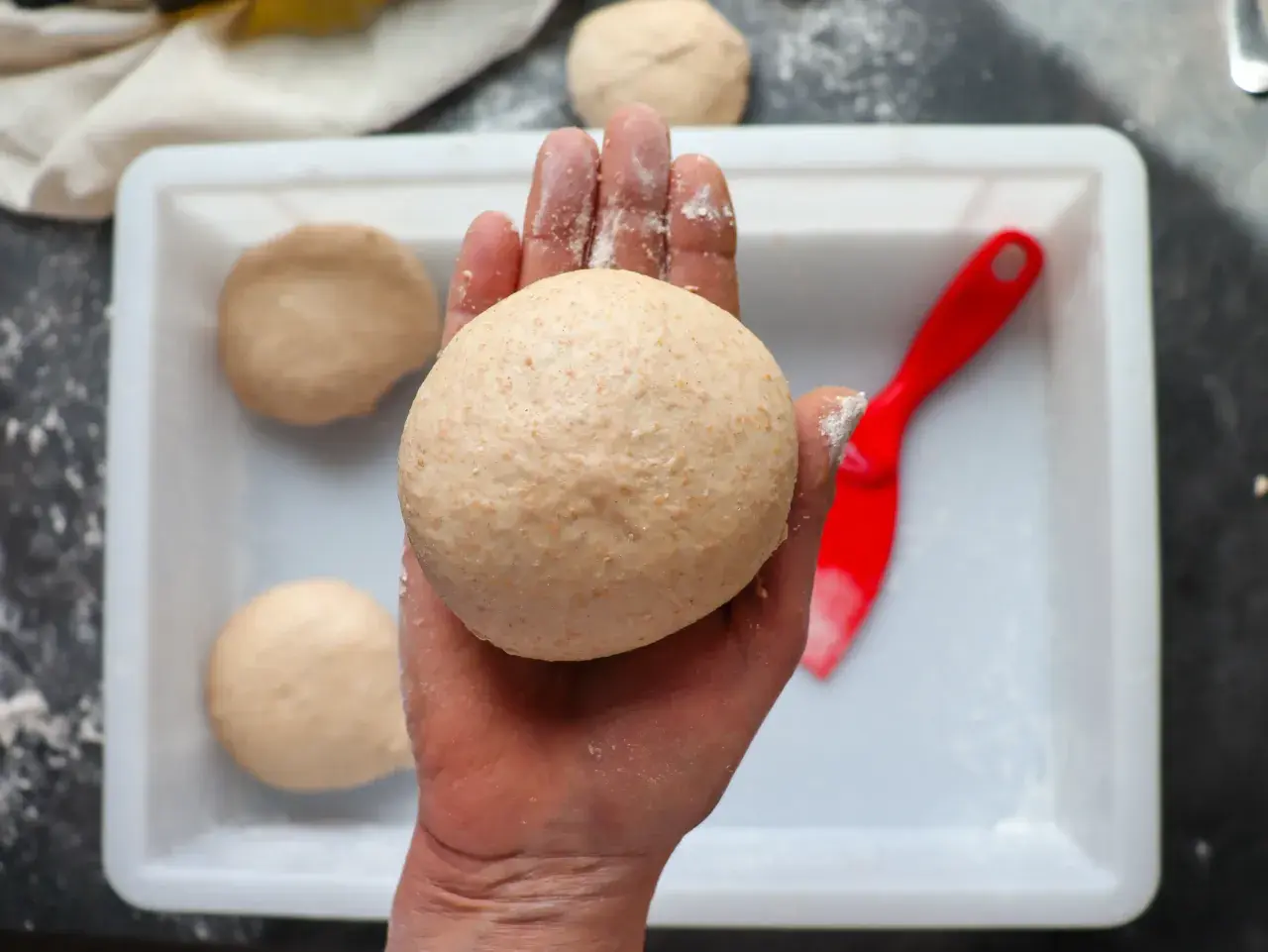 Hand hält klebrigen Pizzateig. Mehr Teiglinge in einer weißen Schale, daneben ein roter Teigschaber.