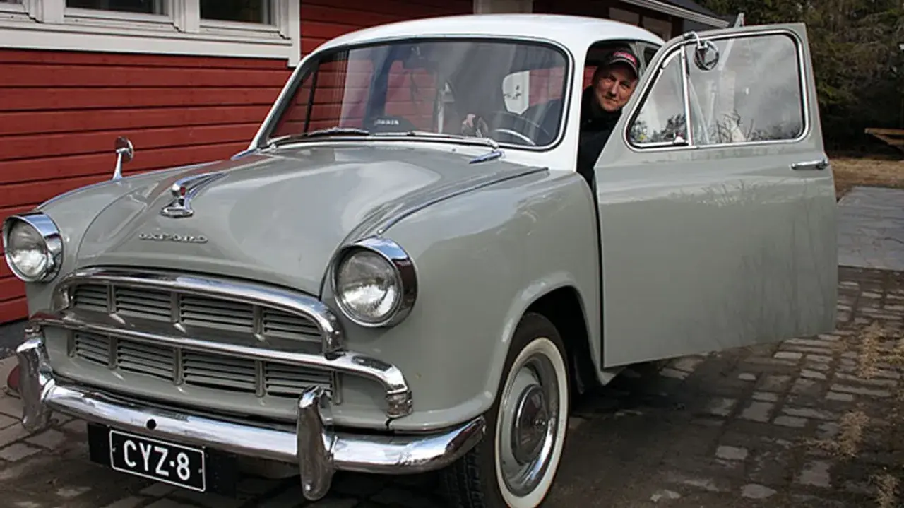 Man ler vid ratten i en grå Morris Oxford med öppen dörr.