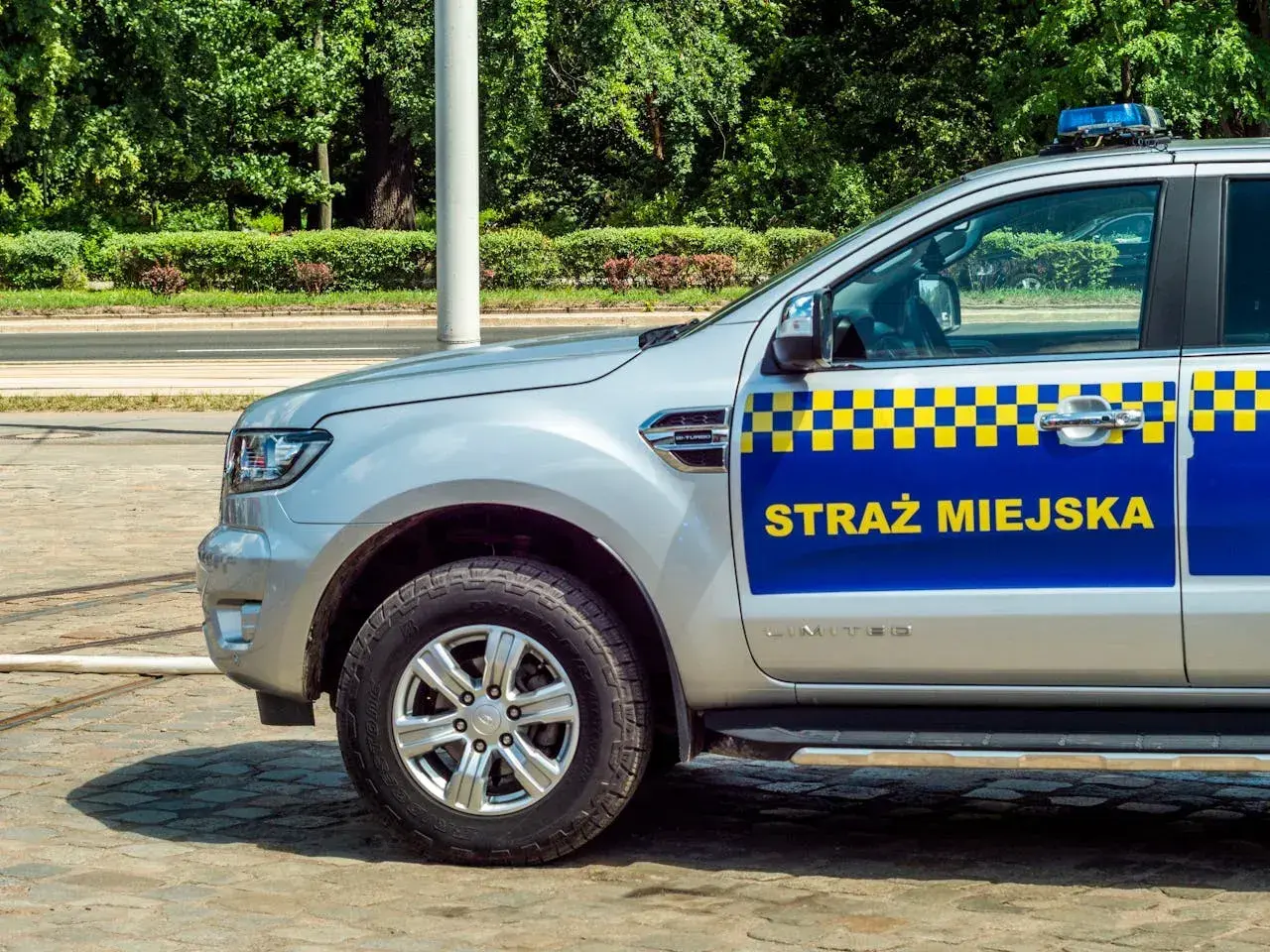 Srebrny samochód Straży Miejskiej z niebiesko-żółtym pasem i napisem "STRAŻ MIEJSKA".